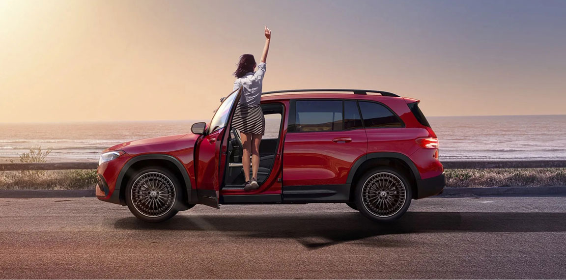 woman standing in red Mercedes-Benz SUV parked by the ocean at sunset