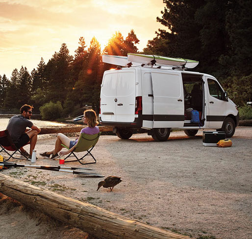 Mercedes-Benz Sprinter Cargo Van parked at a lakeside campsite with two people relaxing in camping chairs at sunset beside the van
