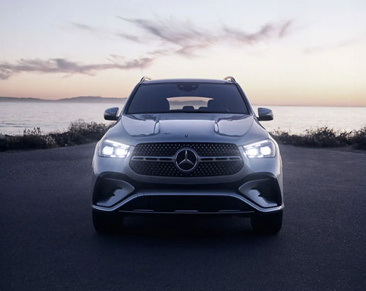 Front view of the Mercedes-Benz GLE SUV with LED headlights illuminated at sunset along a coastal road.