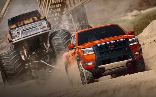 orange Nissan Frontier PRO-4X pickup driving through sand with a monster truck in the background