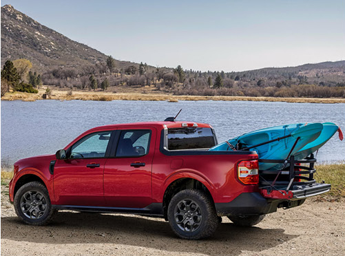 red Ford Maverick pickup truck loaded with kayaks parked beside a lake