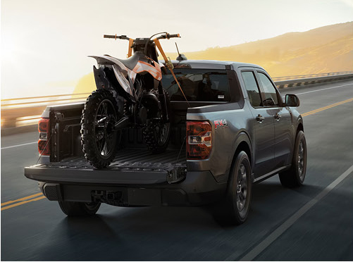 Ford Maverick pickup truck transporting a dirt bike with the tailgate lowered on a highway drive