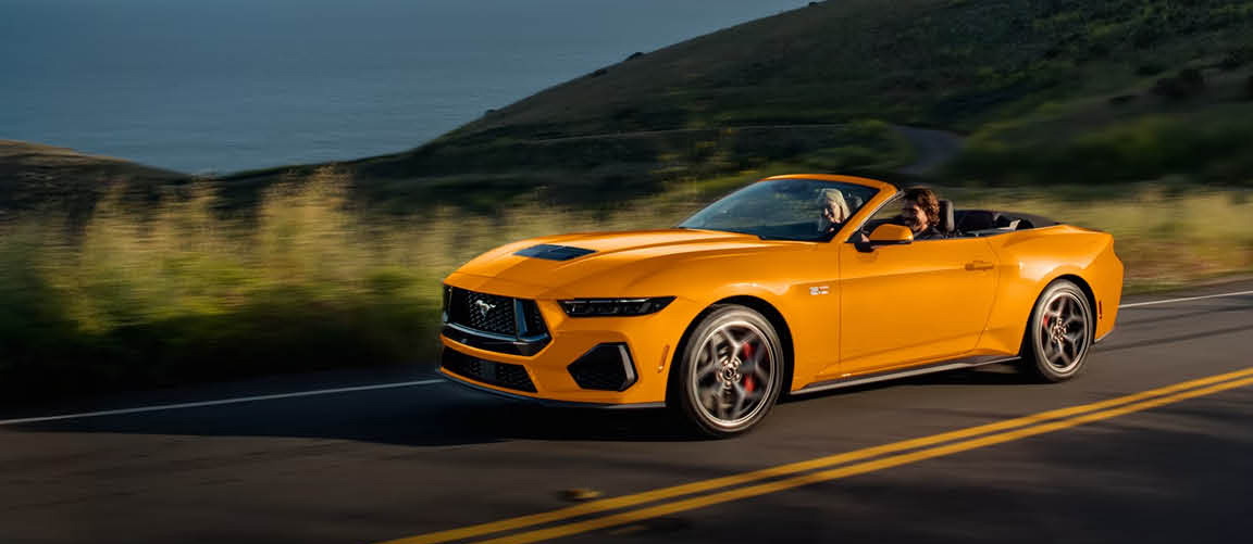 Orange convertible Ford Mustang driving on a scenic coastal road at sunset
